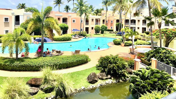 A resort pool surrounded by palm trees and a small pond, with a pathway leading to a building with balconies. - Fairway Courts Studio in Palmas del Mar- Great Pool, Just Mins to Hotel/Beach (Humacao)