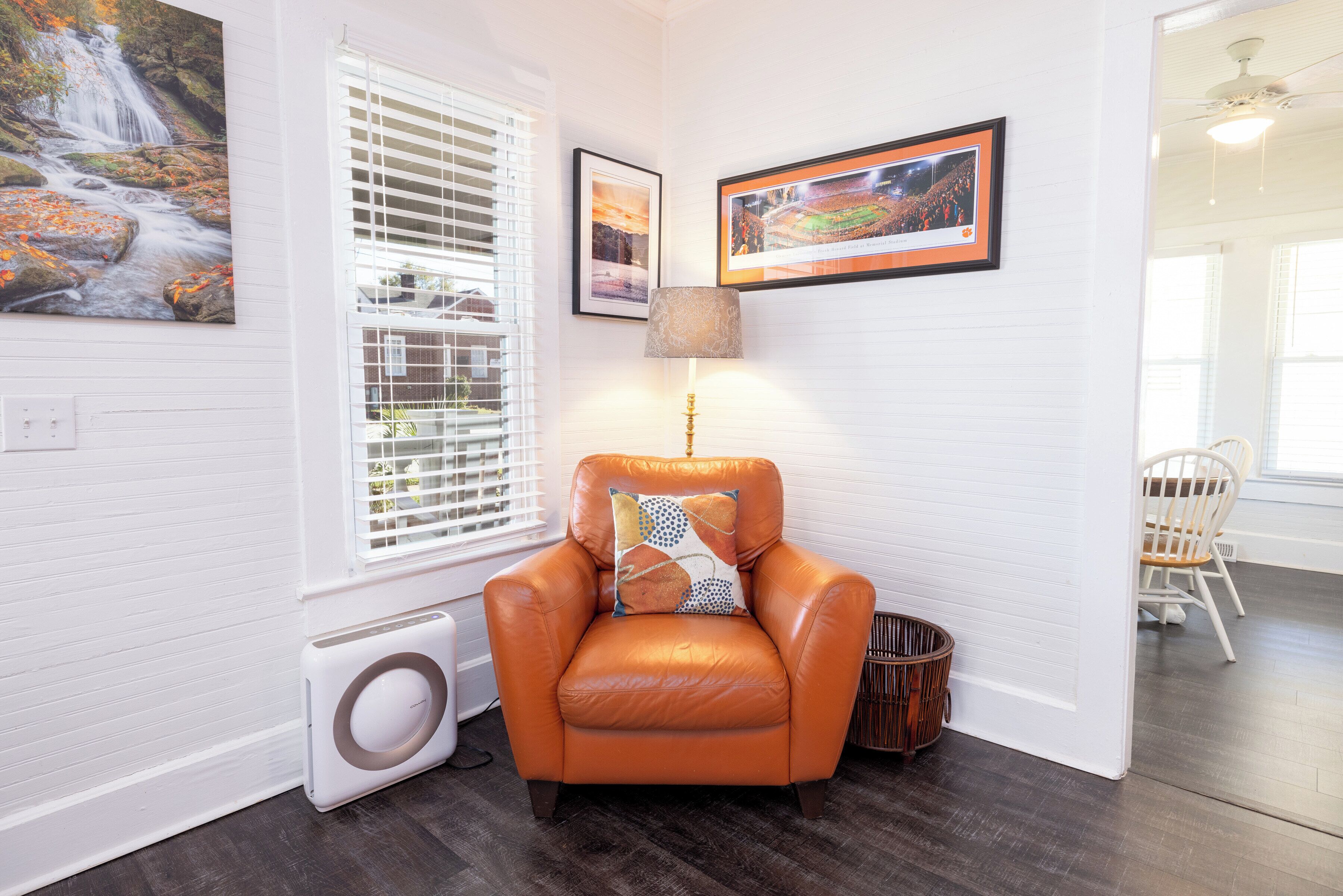 Una habitación con pared blanca, una ventana con persianas, un calentador, un sillón de cuero naranja con cojín estampado, una lámpara y dos cuadros enmarcados.