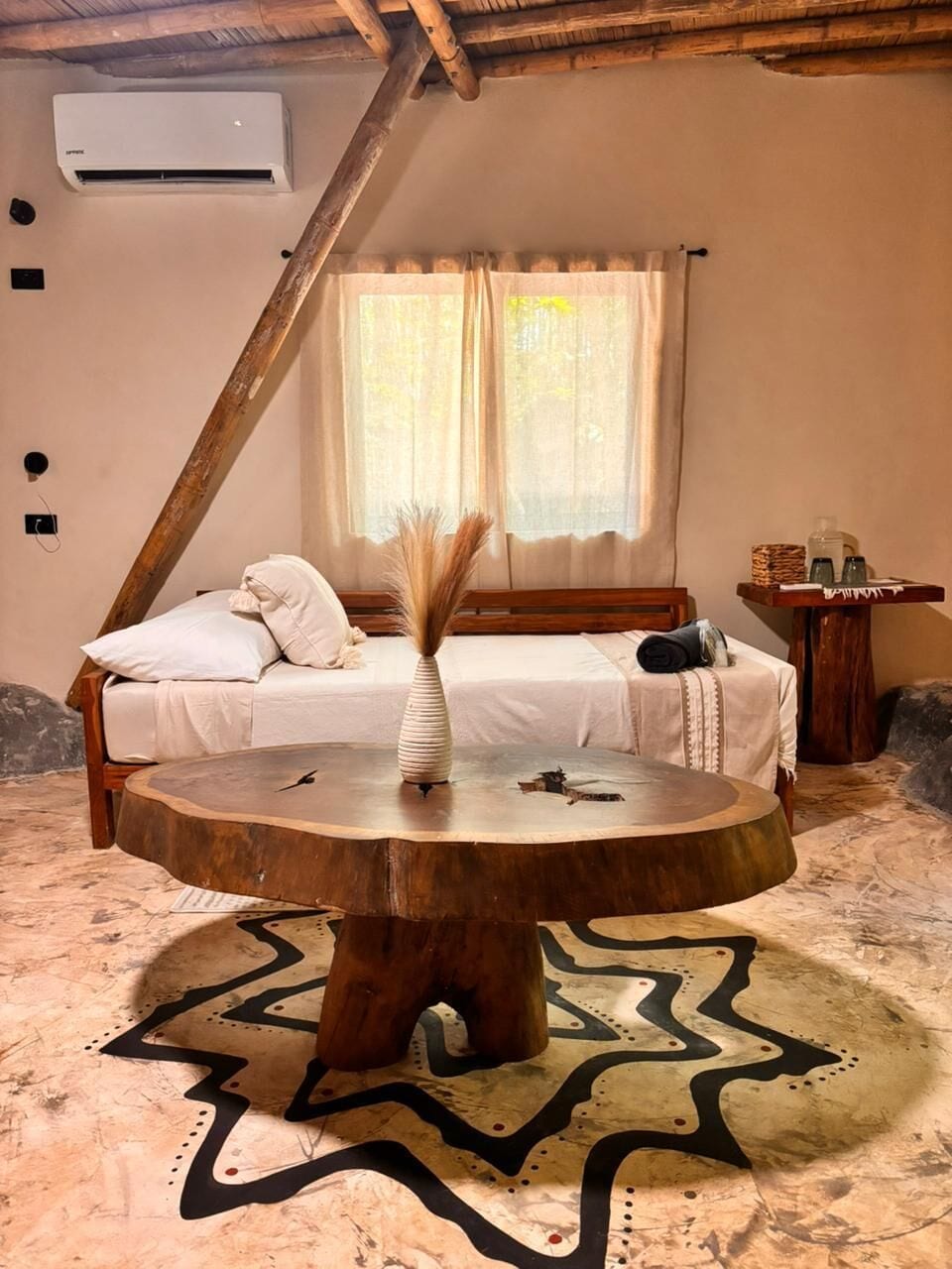 A bedroom with a bed, a bedside table, a round wooden table, and a window with curtains.