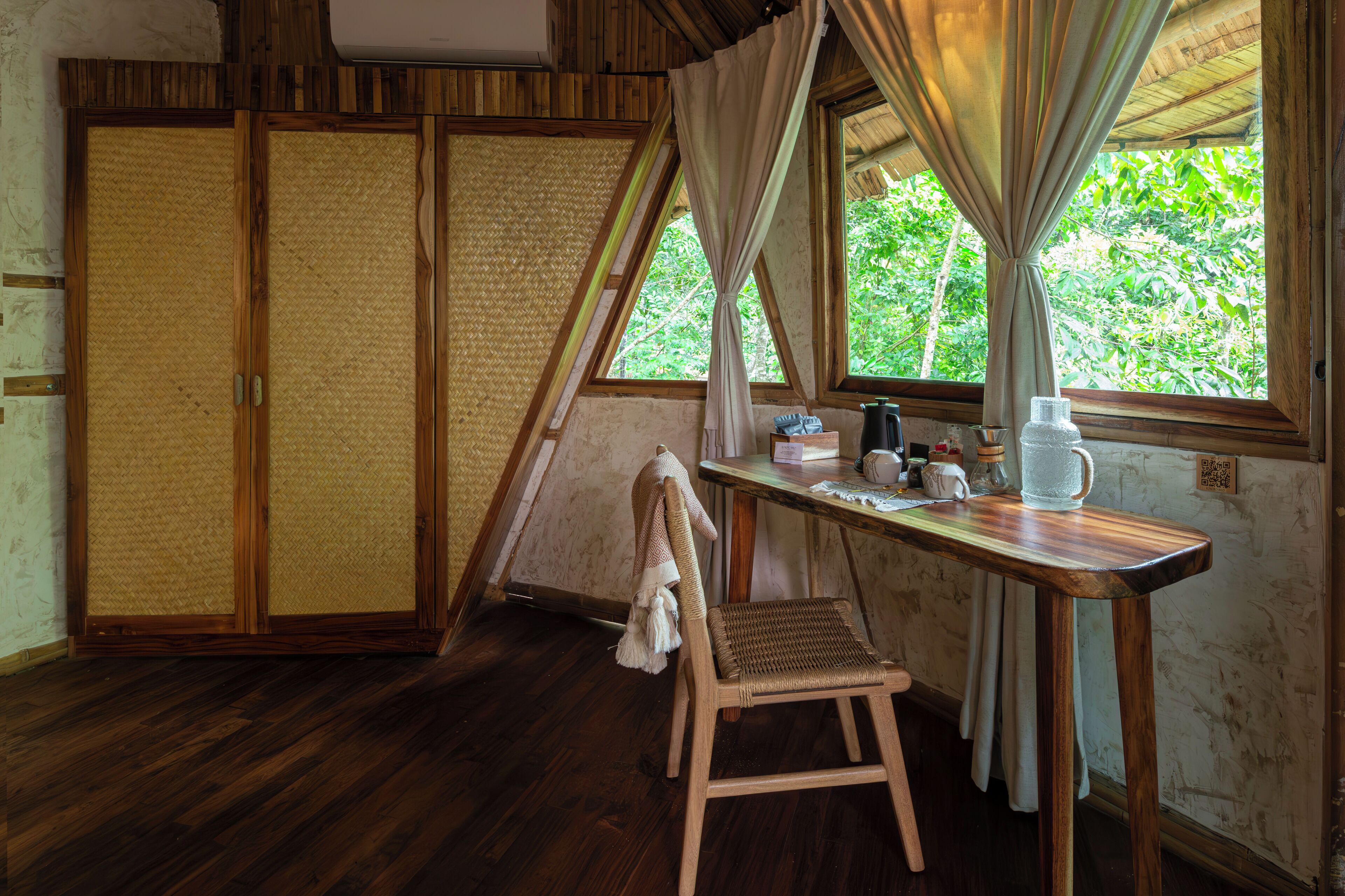 Una habitación con piso de madera, una silla tejida, una mesa con un hervidor y vistas a la vegetación a través de una ventana con cortinas.