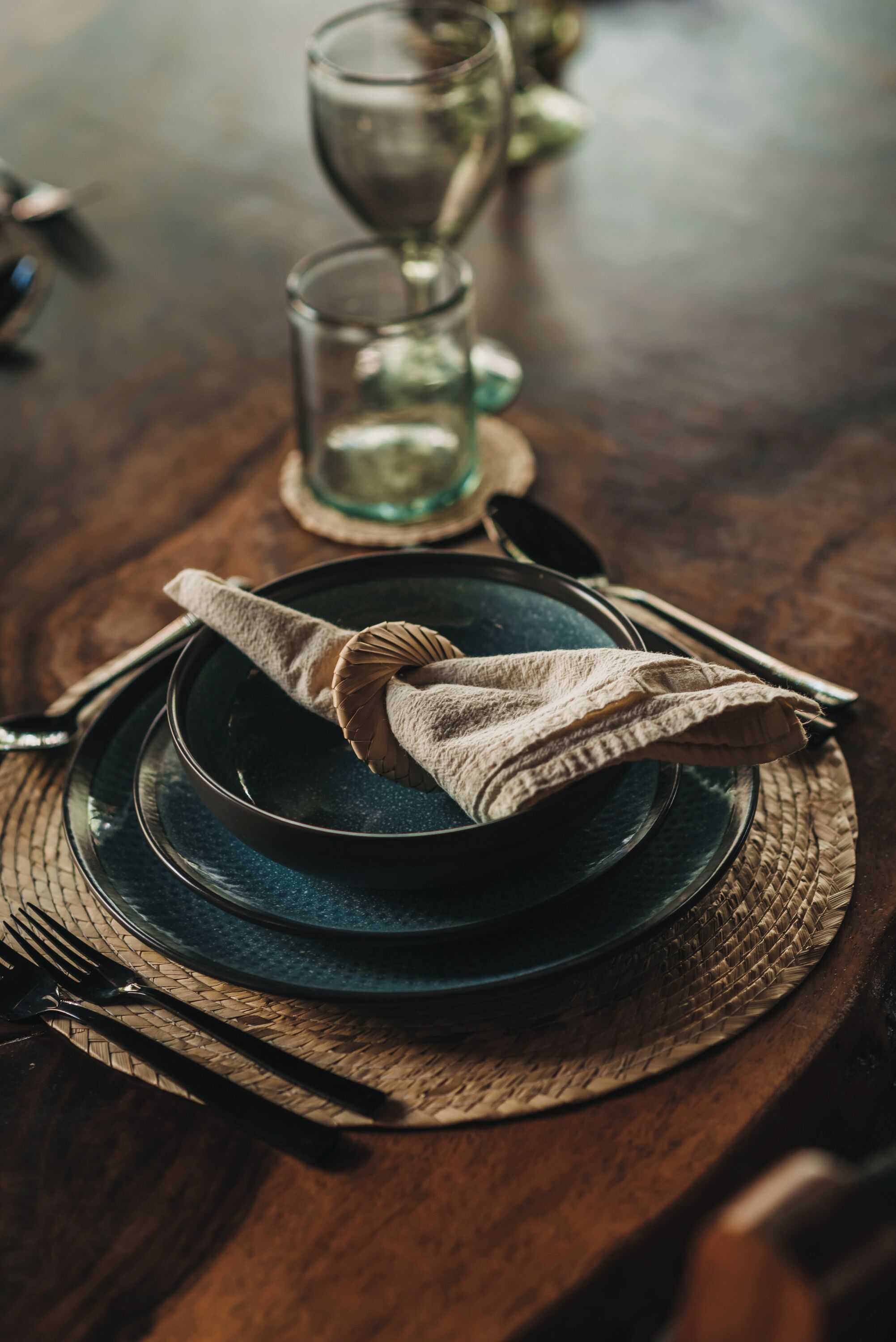 A wooden table set with a dark blue plate, a folded cloth napkin, a fork, and a glass.