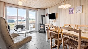 A cozy room with a wooden ceiling, a fireplace, a dining table with chairs, a TV mounted on the wall, and a large window offering a view of the outside. - Beautiful home in Thisted (Thisted)