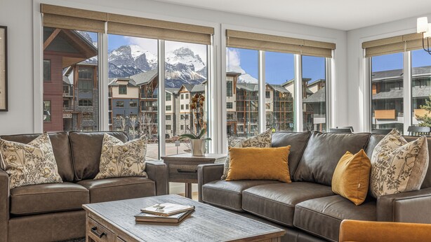 A modern living room with a brown leather sofa, a wooden coffee table, and large windows offering a view of a mountain resort. - Spacious Premier Mountain Retreat conveniently located in Spring Creek! (Canmore)