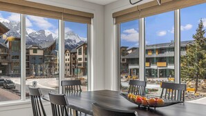 A dining area with a table and chairs, a fruit bowl, and a view of a mountain and buildings. - Spacious Premier Mountain Retreat conveniently located in Spring Creek! (Canmore)