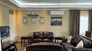 A living room with a brown leather sofa, a brown leather ottoman, a television, and three clocks displaying different cities. - The Residence Inn (Lilongwe)