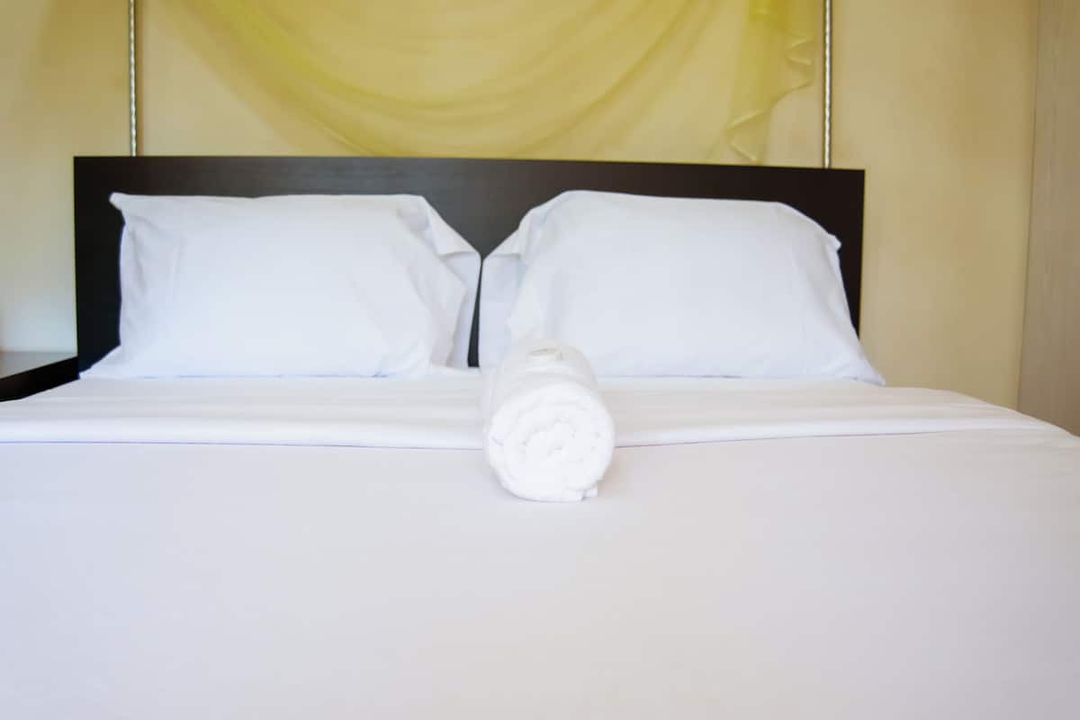 Cama bem arrumada com lençóis e almofadas brancos, uma toalha enrolada no centro e uma cortina amarela ao fundo.