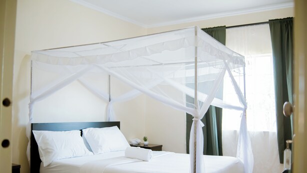 A four-poster bed with a canopy, a bedside table with a lamp, and a window with curtains. - The Residence Inn (Lilongwe)