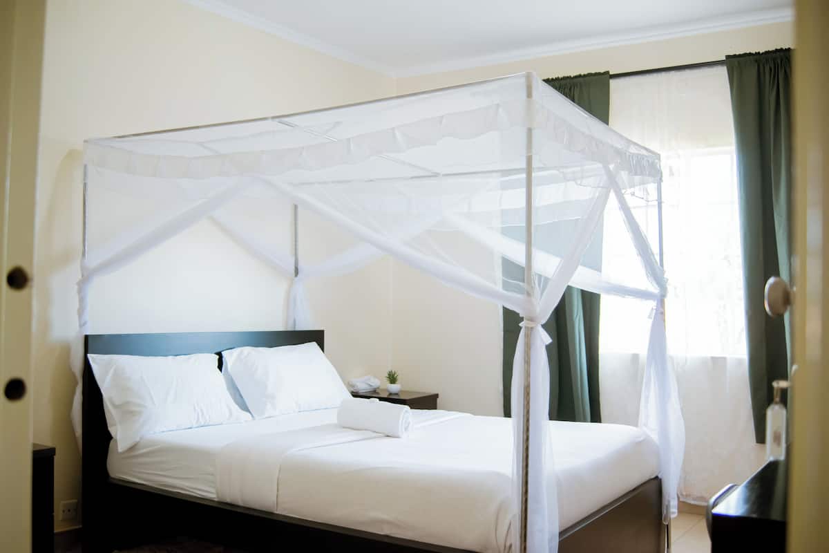 Uma cama com dossel e cortinas, uma mesa de cabeceira com abajur, e uma janela com cortinas.