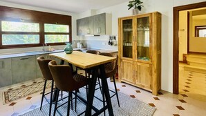 Une cuisine moderne avec une table en bois, des tabourets de bar, un meuble avec des portes en verre et une fenêtre donnant sur un paysage verdoyant.