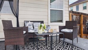 A covered patio with wicker furniture, a patterned rug, and a window with curtains.