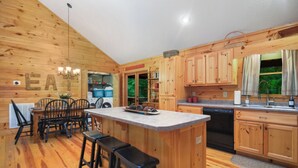 A wooden cabin-style kitchen with a central island, bar stools, a dining table with chairs, and a view of the outdoors. - Serenity Moon, a Spacious Cabin in Coosawattee River Resort, North GA Mountains (Ellijay)
