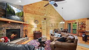 Un intérieur de chalet chaleureux avec une cheminée en pierre, un canapé en cuir, un coin repas et un ventilateur de plafond.