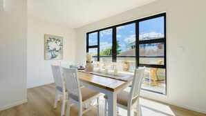 Un espace repas avec une table en bois, des chaises blanches et une grande fenêtre donnant sur des immeubles et un ciel dégagé.
