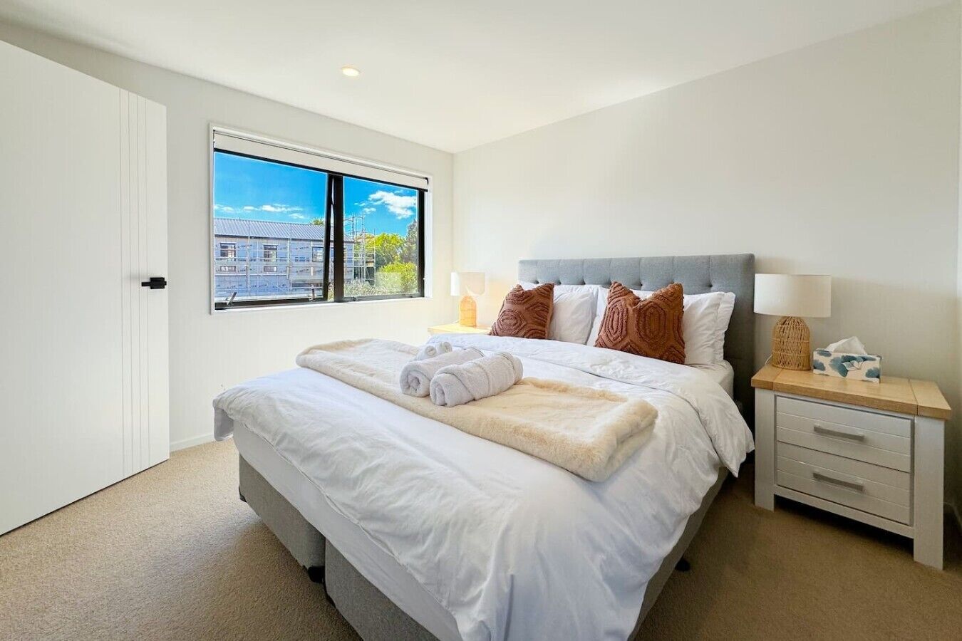 A bedroom with a large bed, a nightstand, a window with a city view, and a door.