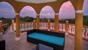 A covered outdoor seating area with a sofa and a table, surrounded by arches and a railing, overlooking a scenic view.