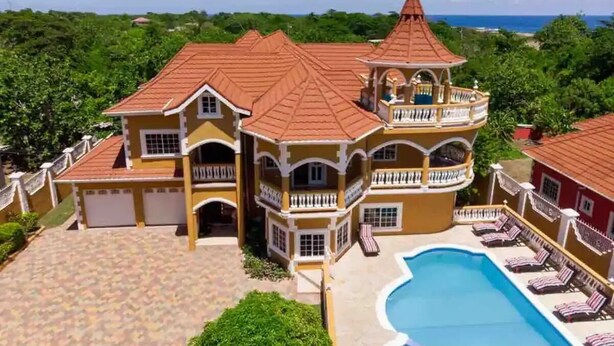 A large, multi-story house with a swimming pool, surrounded by lush greenery and a clear view of the ocean.