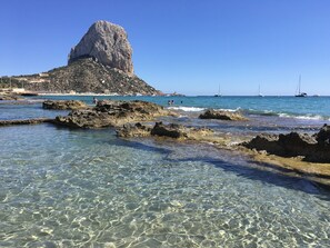 A rocky shoreline with clear water, a prominent rock formation, and boats in the distance.