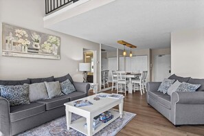 Un salon moderne avec deux canapés gris, une table basse blanche et un coin repas comprenant des chaises blanches et une table en bois.