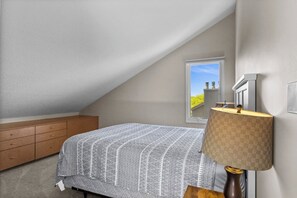 A bedroom with a sloped ceiling, a window with a view, a wooden dresser, and a lamp. - Traverse city/Golf/Hiking/Equestrian/Lake (Williamsburg)