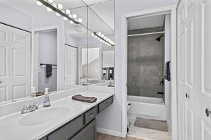 A modern bathroom with a large mirror, two sinks, a shower, and a walk-in closet.