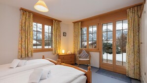 Ein Hotelzimmer mit zwei Betten, einem Sessel, einem Nachttisch mit Lampe und großen Fenstern mit Vorhängen.