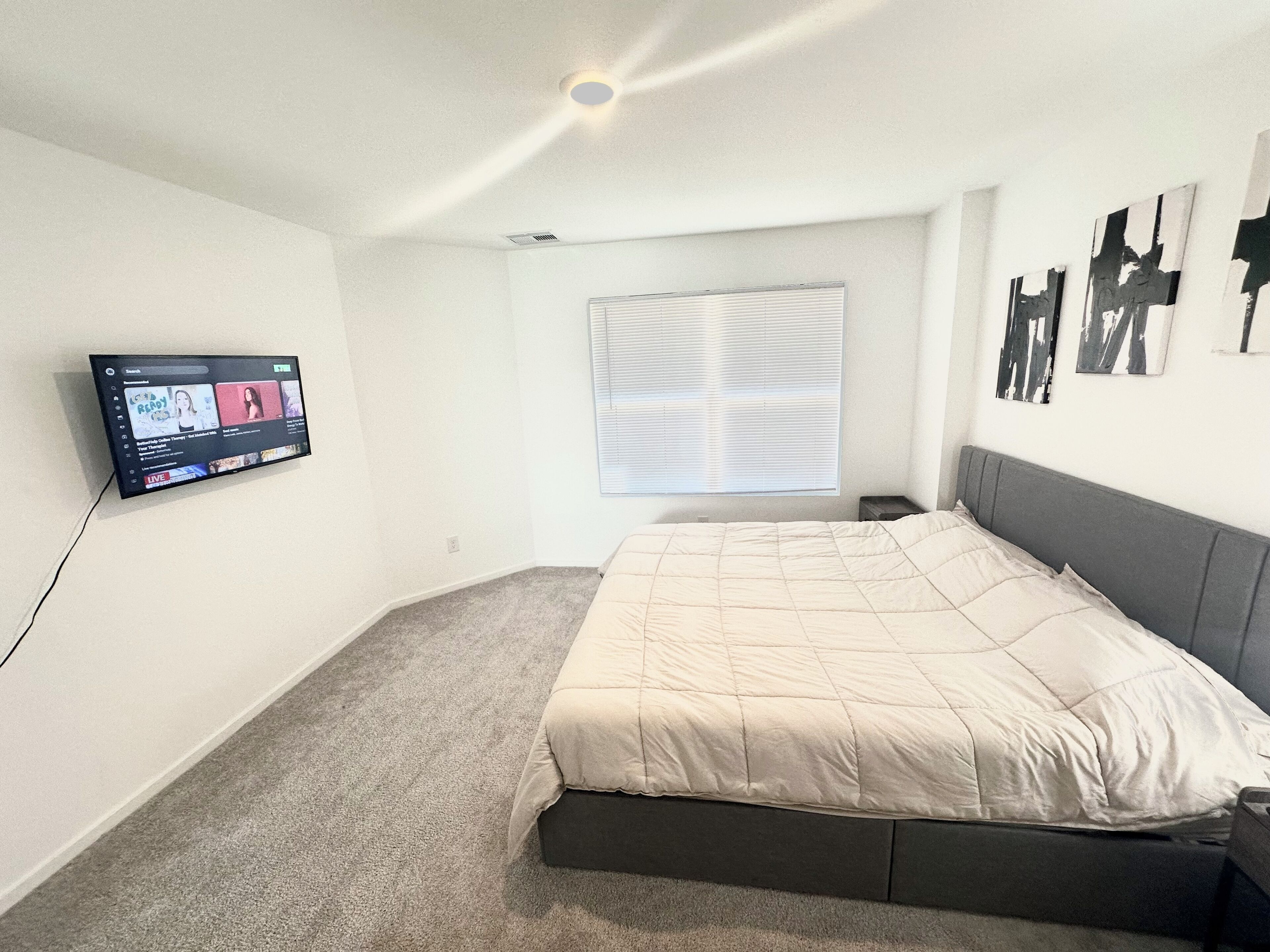 Ein Schlafzimmer mit einem Bett, einem an der Wand montierten Fernseher, einem Fenster mit Jalousien und einem kleinen Nachttisch.