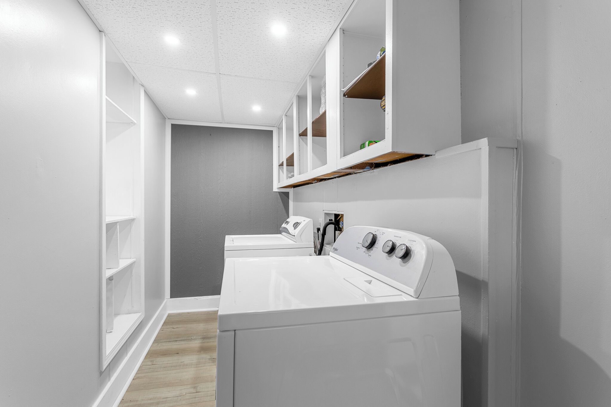 A laundry room with two white washers and dryers, wooden shelves above the dryers, and a grey wall.