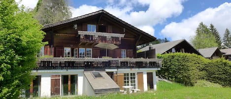 A two-story wooden chalet with a balcony, a small attached structure with a table and chairs, surrounded by a grassy field and trees.