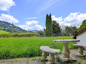 A garden with a stone table and bench, surrounded by green grass and flowers, with mountains in the background.