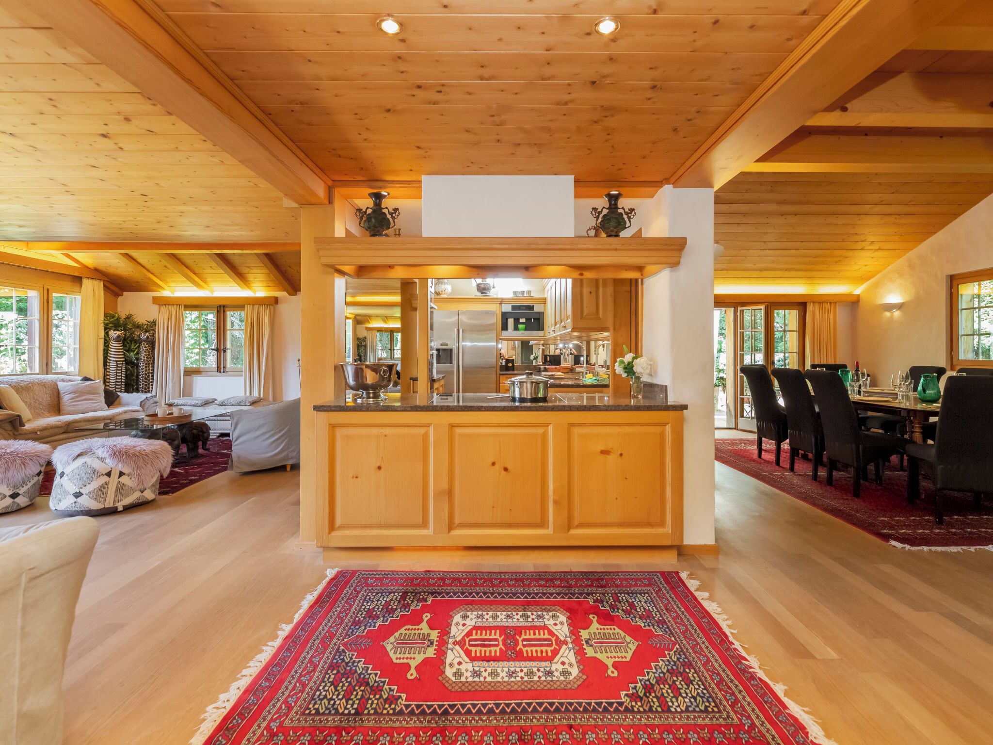 A spacious living area with a wooden ceiling, a kitchen with a central island, and a dining area with a red patterned rug.