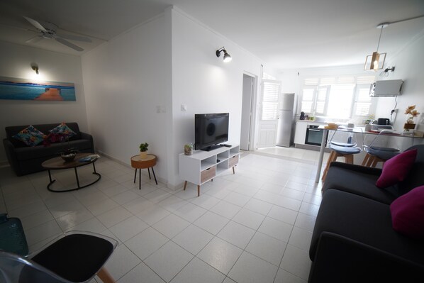 A modern living room with a flat-screen TV, a sofa, a coffee table, and a dining area with a kitchen in the background. - Eden T2 Apartment, 200 meters from the beach—ideal for families (Sainte-Luce)