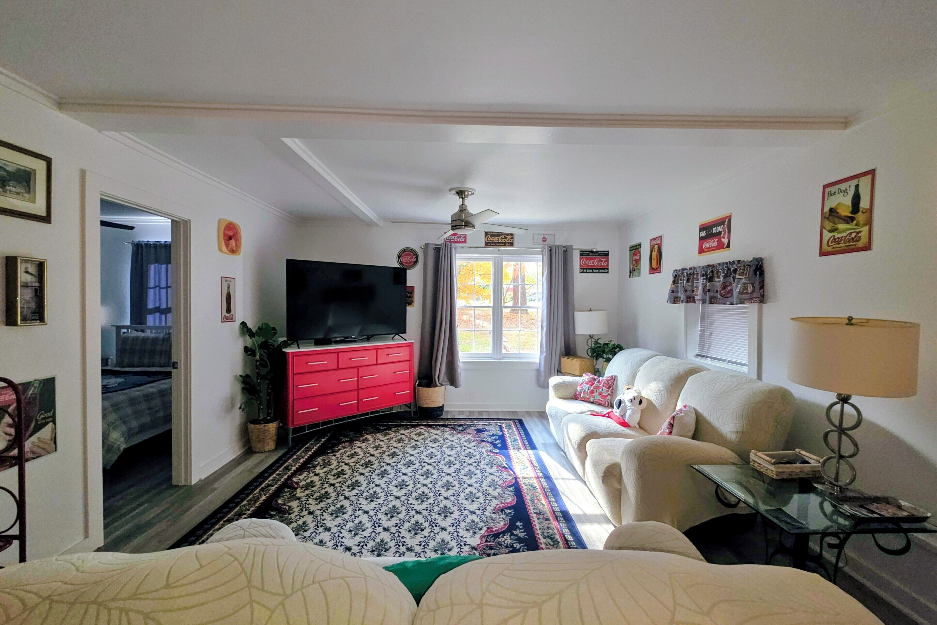 Ein Wohnzimmer mit Fernseher, rotem Schminktisch, weißem Sofa und einem gemusterten Teppich.
