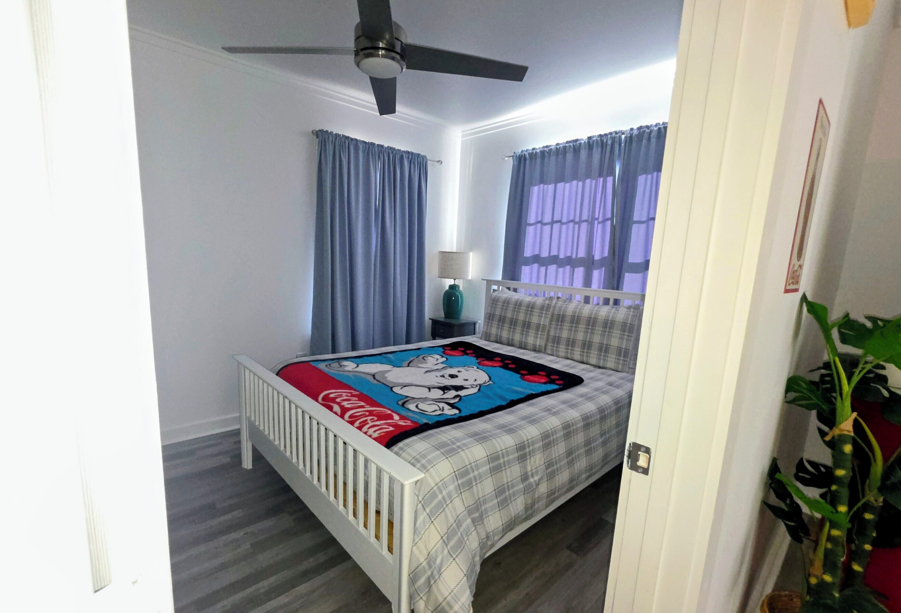 Une chambre à coucher avec un lit, un ventilateur de plafond, des rideaux, une table de chevet, une lampe et une plante.