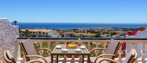 Une terrasse dotée de meubles en osier et d’une table dressée pour le repas, offrant une vue sur une ville côtière et la mer.