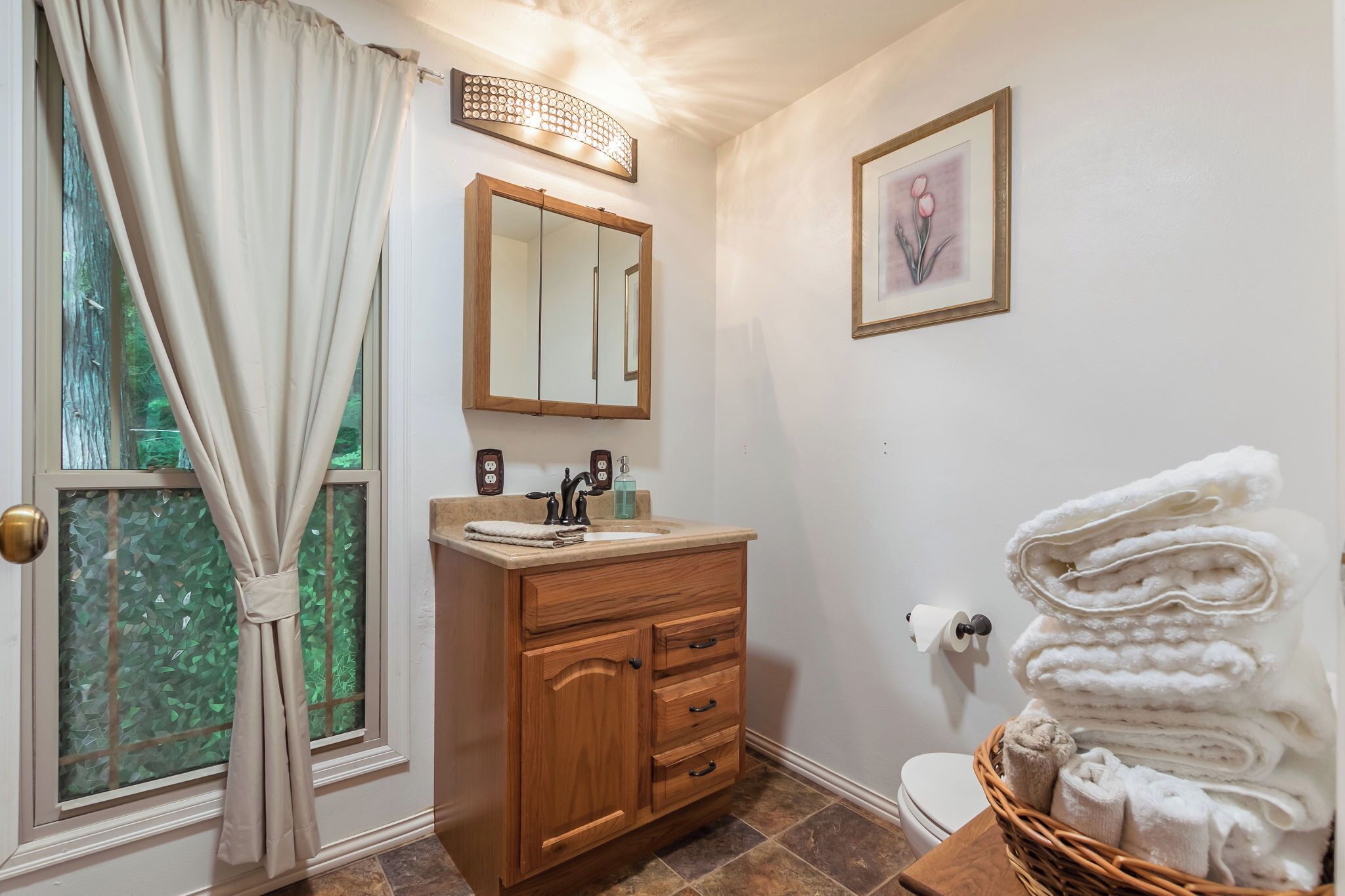 Une salle de bain avec un meuble-lavabo en bois, un évier, un miroir, des toilettes et une fenêtre avec des rideaux.