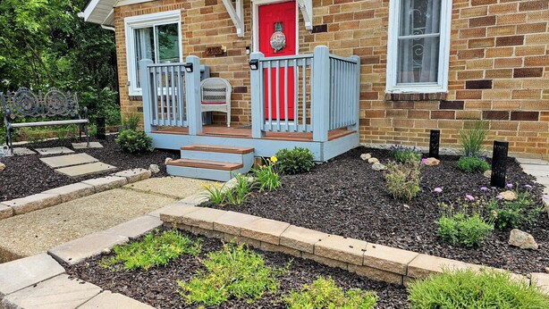 A brick house with a red door, a porch with a blue railing, and a garden with stepping stones and plants.