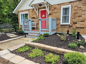 Une maison en briques avec une porte rouge, un porche doté d’une balustrade bleue et un jardin agrémenté de pas japonais et de plantes.
