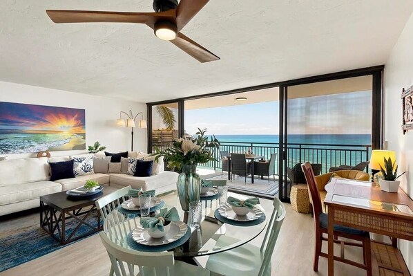 A modern living room with a glass dining table, a sofa, and a large painting of a sunset.