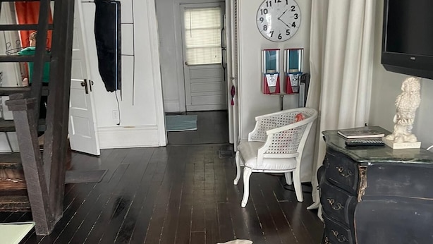 A room with dark wooden flooring, a white door, a clock, a white chair, a black dresser, and a television. - Contemporary tiny home with loft bedroom in the Irish Channel, private courtyard (New orleans)