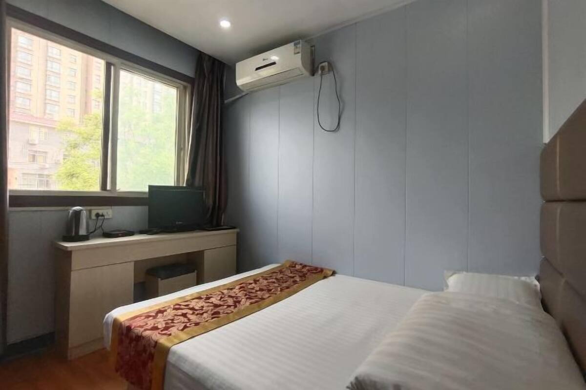 A hotel room with a bed, a desk with a computer, an air conditioning unit, and a window with curtains.