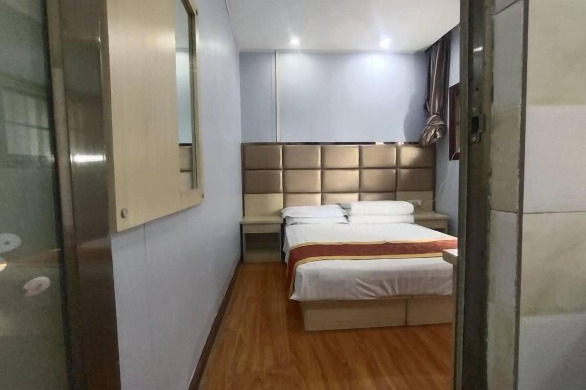 A narrow hotel room with a bed, a nightstand, a wall-mounted headboard, and a small window.