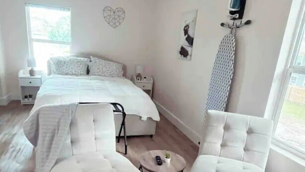 A modern bedroom with a bed, two white armchairs, a round coffee table, and a TV mounted on the wall. - Sun & Speedway Villa - Centrally located to everything. (Daytona Beach)