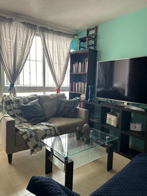 A living room with a sofa, a glass coffee table, a bookshelf, a television, and a window with curtains.