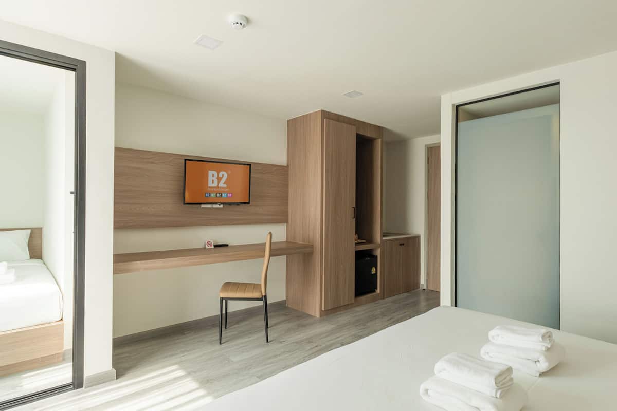 A modern hotel room with a bed, a desk, a chair, a television, and a wardrobe.