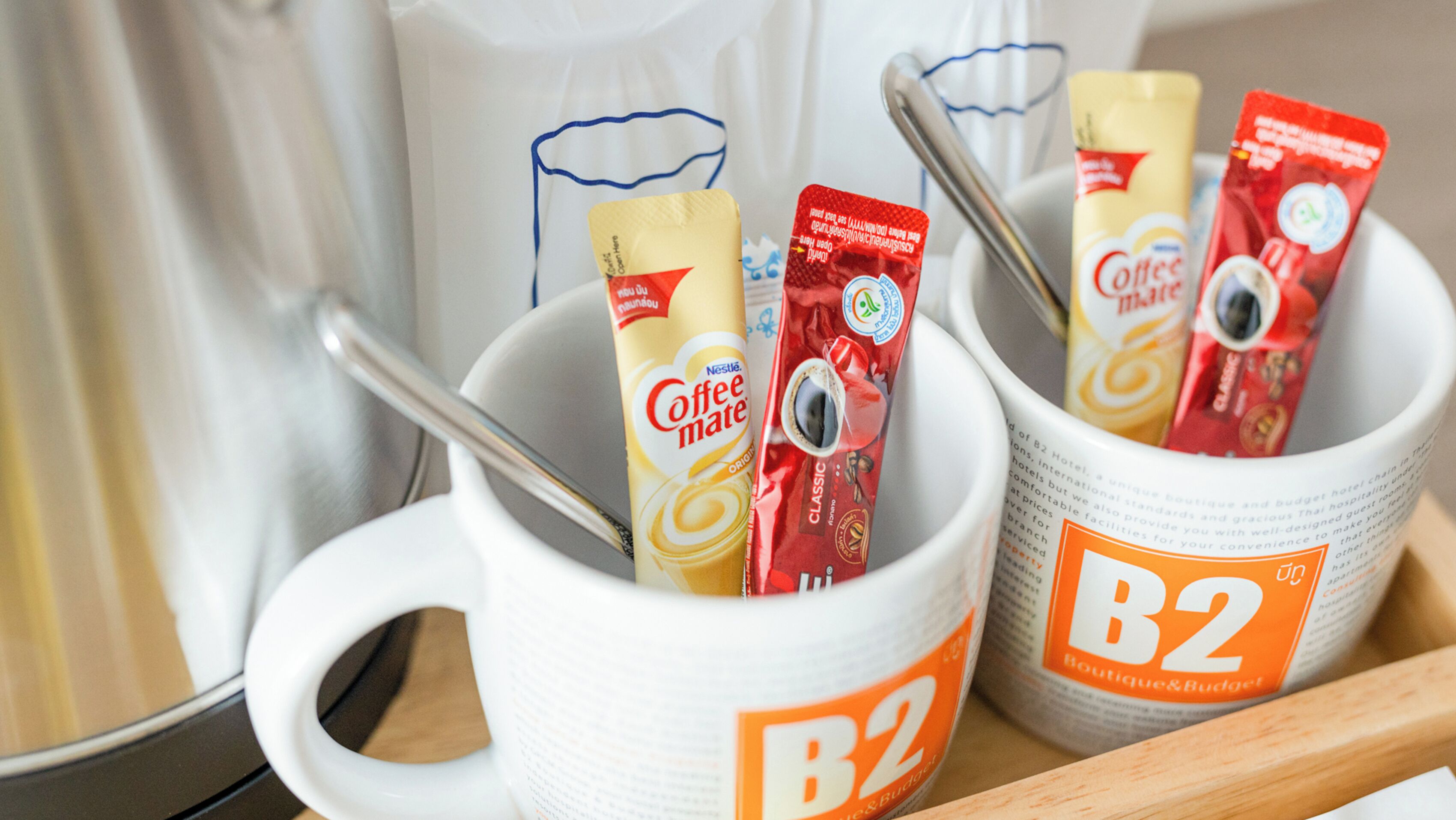 Två vita muggar med B2 Boutique & Budget-logotyp, var och en innehåller en påse kaffe och en sked.