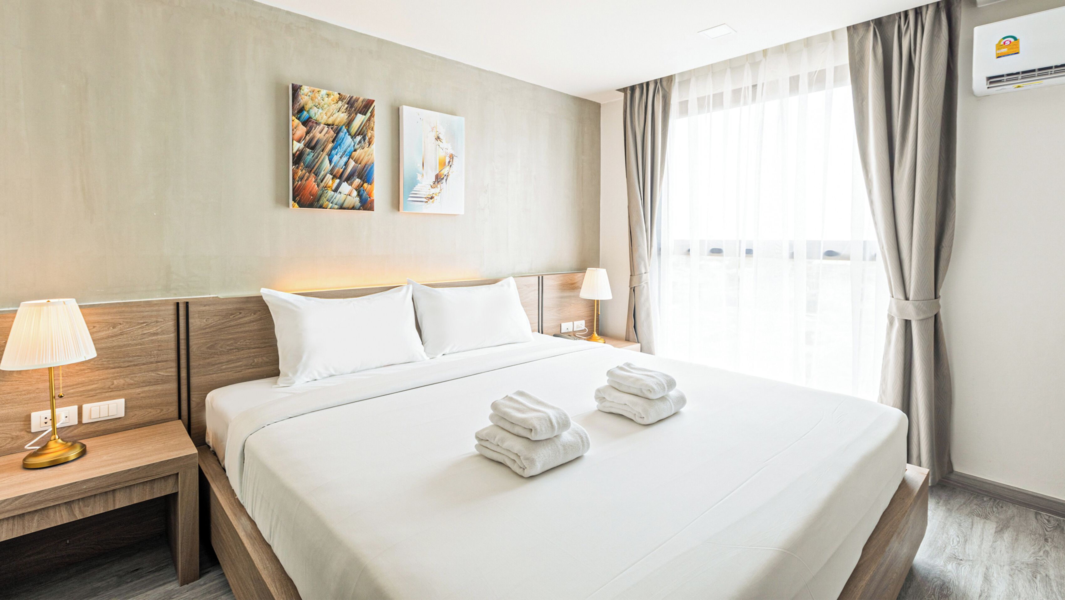 Een hotelkamer met een groot bed, twee nachtkastjes, een lamp, een wandairconditioner en een raam met gordijnen.