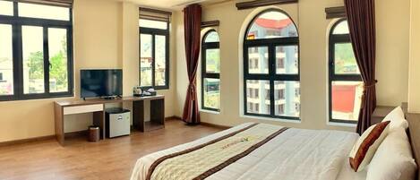 Habitación de hotel con una cama grande, un escritorio, televisor y ventanas amplias con cortinas.