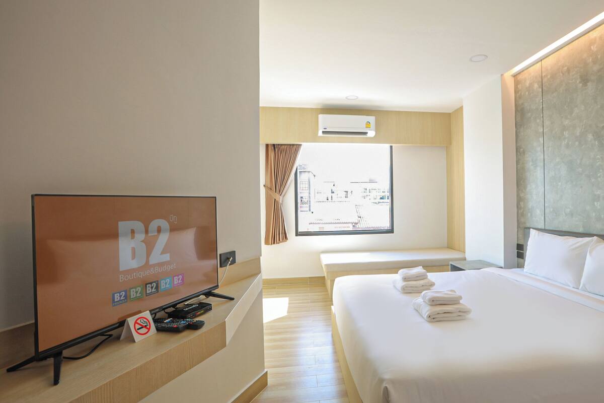 Habitación de hotel con una cama grande, un televisor en un soporte, una ventana con cortinas y vistas al exterior.