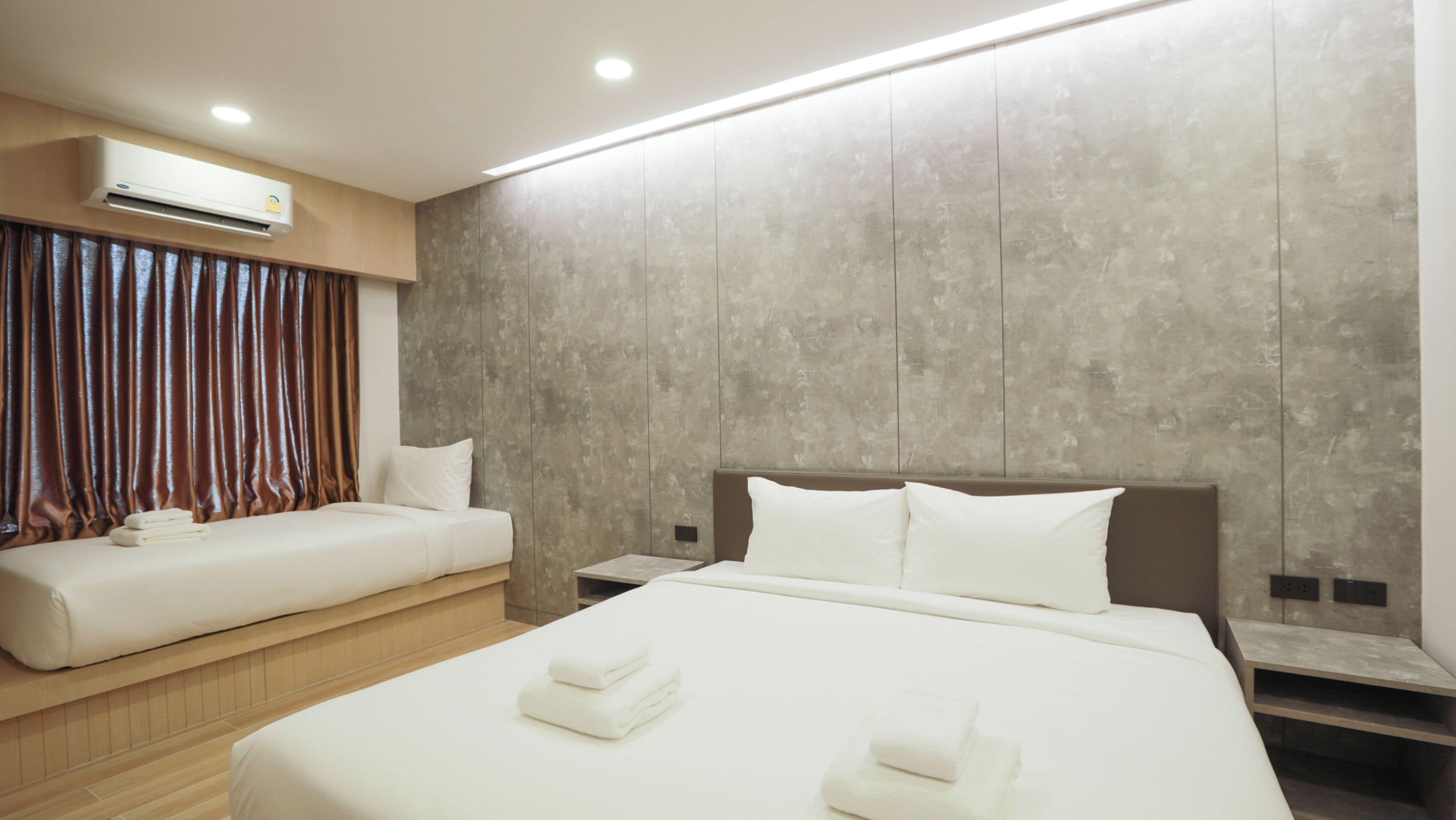 A hotel room with two beds, a wooden floor, a large window with curtains, and an air conditioning unit on the wall.