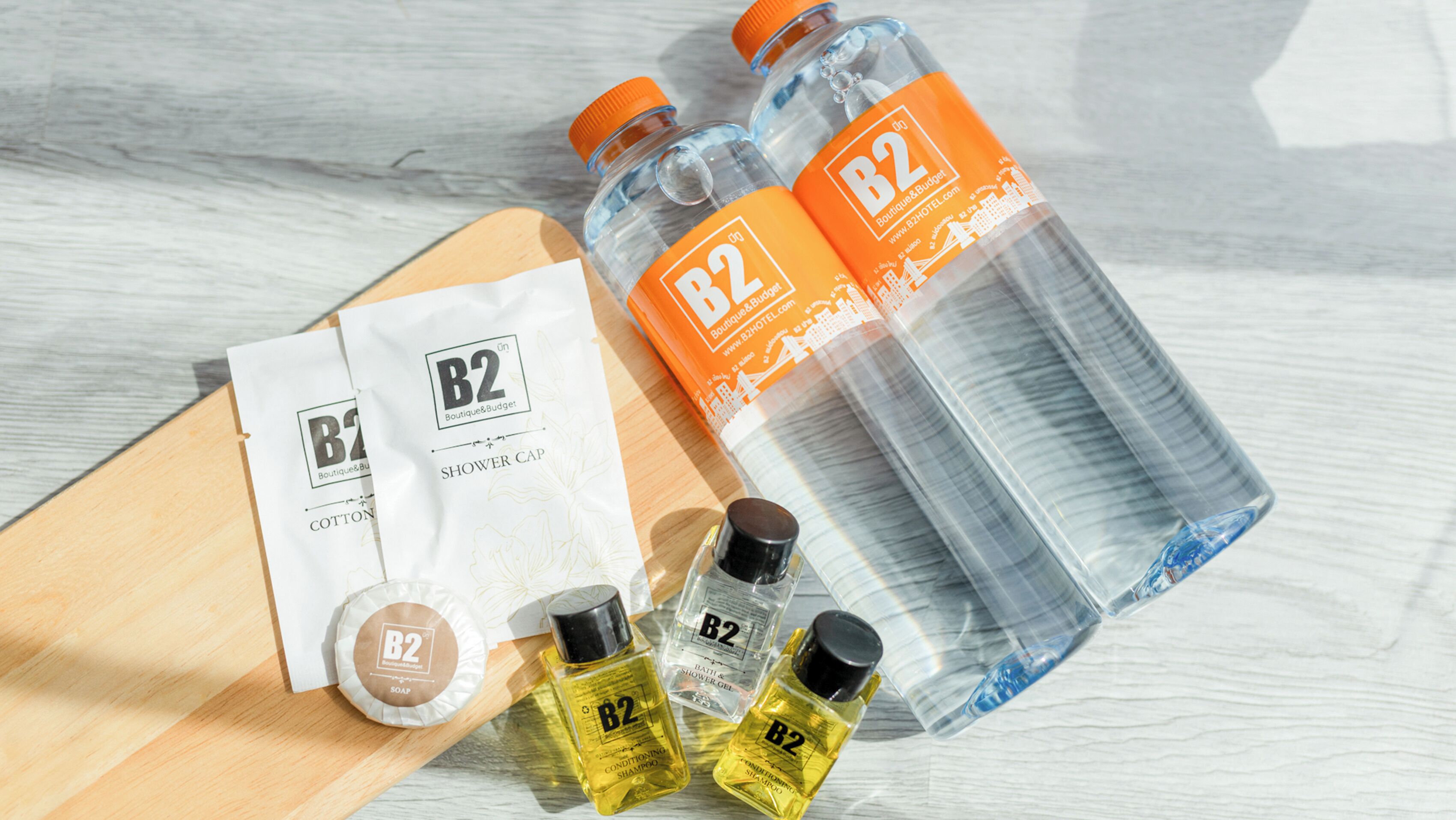 A wooden surface with various B2 branded toiletries including a shower cap, cotton pad, soap, and conditioning shampoo.
